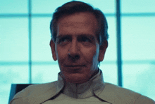 a man in a white jacket looks at the camera with a window in the background