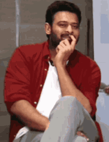a man in a red shirt and white shirt is sitting with his legs crossed and his hand on his mouth .