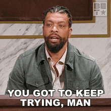 a man with a beard is sitting in front of a sign that says " you got to keep trying man "