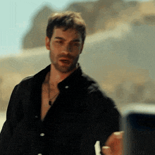 a man with a beard wearing a black shirt and a necklace is standing in front of a mountain .