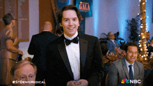 a man in a tuxedo and bow tie smiles in front of a sign that says gala