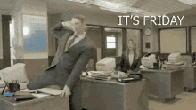 a man in a suit and tie is sitting on a desk in an office with the words it 's friday behind him
