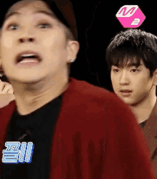 a man in a red jacket is standing next to another man with a m2 logo on his head ..