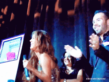 a woman laughs while a man and woman applaud in front of a screen that says anaioverdose on the bottom