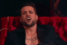 a man with a tattoo on his chest is sitting on a red couch and smiling .