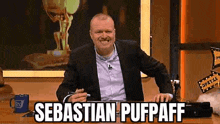 a man in a suit is sitting at a table with the words sebastian pufpaff written on the screen .
