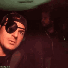two men are sitting next to each other in a dark room . one of the men is wearing a pirate eye patch .
