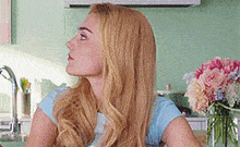 a woman with long blonde hair is sitting in a kitchen with a vase of flowers .
