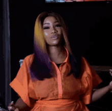 a woman with purple and yellow hair is wearing an orange shirt and sitting in front of a television .