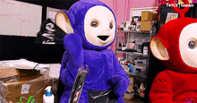 a purple teletubbies mascot is holding a pen in a room with other stuffed animals