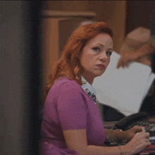 a woman with red hair is sitting at a desk in front of a phone