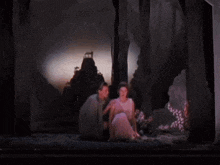 a man and woman are sitting in a dark forest with a mountain in the background