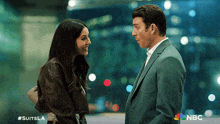 a man in a suit and a woman in a leather jacket are looking at each other with the nbc logo in the background