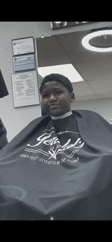 a boy is getting his hair cut by a barber wearing a cape that says greatest stylists in the world
