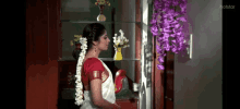 a woman in a red top and white saree is standing in front of a shelf with purple flowers on it .