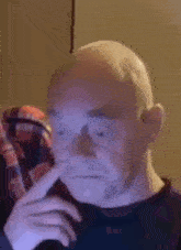 a bald man wearing glasses and a plaid shirt is holding his hand to his nose .