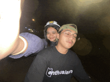 a man wearing a black shirt that says enthusiasm stands next to a woman wearing a helmet