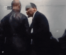 a man in a suit and tie is talking to another man in a dark room with a 24 aok sign in the background .