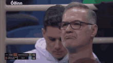 a man wearing glasses is watching a basketball game with a scoreboard behind him that says odinn