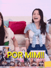 two women are sitting on a couch with a sign that says por mim