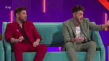 two men are sitting on a blue couch talking to each other .