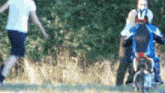 a man is riding a motorcycle in a field while another man stands behind him .