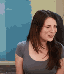 a woman in a grey shirt is smiling in front of a blue wall with azaaarmi written below her