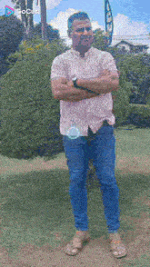 a man in a striped shirt and blue jeans is standing with his arms crossed in a park .
