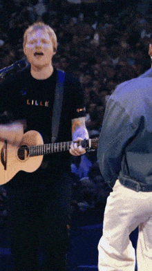 a man singing into a microphone while holding a guitar and wearing a shirt that says lille