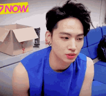 a man in a blue tank top is sitting on a blue couch in front of a now sign