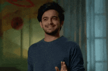 a man with a beard is wearing a blue sweater and smiling while standing in front of a window .