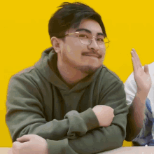 a man wearing glasses and a green hoodie is sitting at a table with his arms crossed