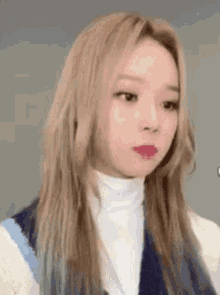 a close up of a woman with long blonde hair wearing a white turtleneck and blue vest .