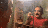 a woman in a red shirt is looking at herself in a bathroom mirror with the letter x in the corner