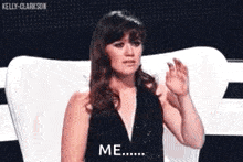 a woman in a black dress is sitting in a chair with her hand up and says `` me '' .