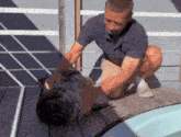 a man in a blue shirt is kneeling on a wooden deck next to a pool