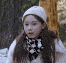 a girl wearing a white hat and a checkered scarf is standing in the woods .