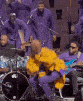 a man in a yellow jacket is sitting on a stool on a stage playing drums .