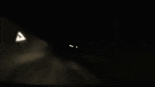 a black and white photo of a car driving down a dark road at night .
