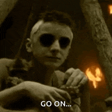 a man with glowing eyes is sitting in a dark room with a fire in the background and says `` go on '' .