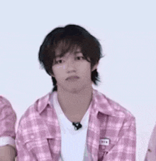 a young man wearing a pink plaid shirt and a white shirt is sitting in front of a white wall .