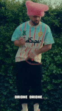 a man wearing a pink hat and a striped shirt is standing in front of a hedge .