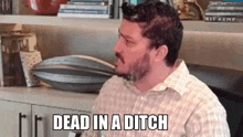 a man with a beard is sitting in a chair in front of a bookshelf and says dead in a ditch .