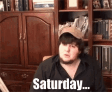 a man with a beard and hat is sitting in front of a bookshelf and says saturday .