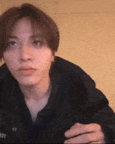 a young man in a black jacket is looking at the camera while sitting in front of a wall .