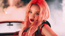 a woman with pink hair and red lips is sitting in a red car and looking at the camera .