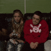 a man wearing a red sweater with a skull and crossbones on it sits next to a woman holding a video game controller
