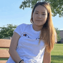 a young woman is sitting on a bench in a park wearing a white champion t-shirt .