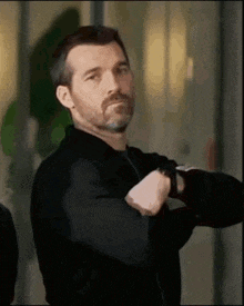 a man with a beard wearing a black jacket and a watch is looking at his watch .
