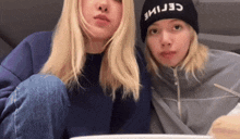 a woman wearing a celine hat sits next to a woman wearing a blue sweater